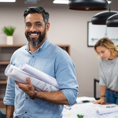 Portrait of multiethnic architect with blueprints in creative office. Mature middle eastern contractor holding roll of architectural projects while looking at camera. Happy latin man in casual standing in meeting room with engineers working in background.