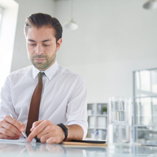 Young confident lawyer making working notes on paper while sitting by desk in office